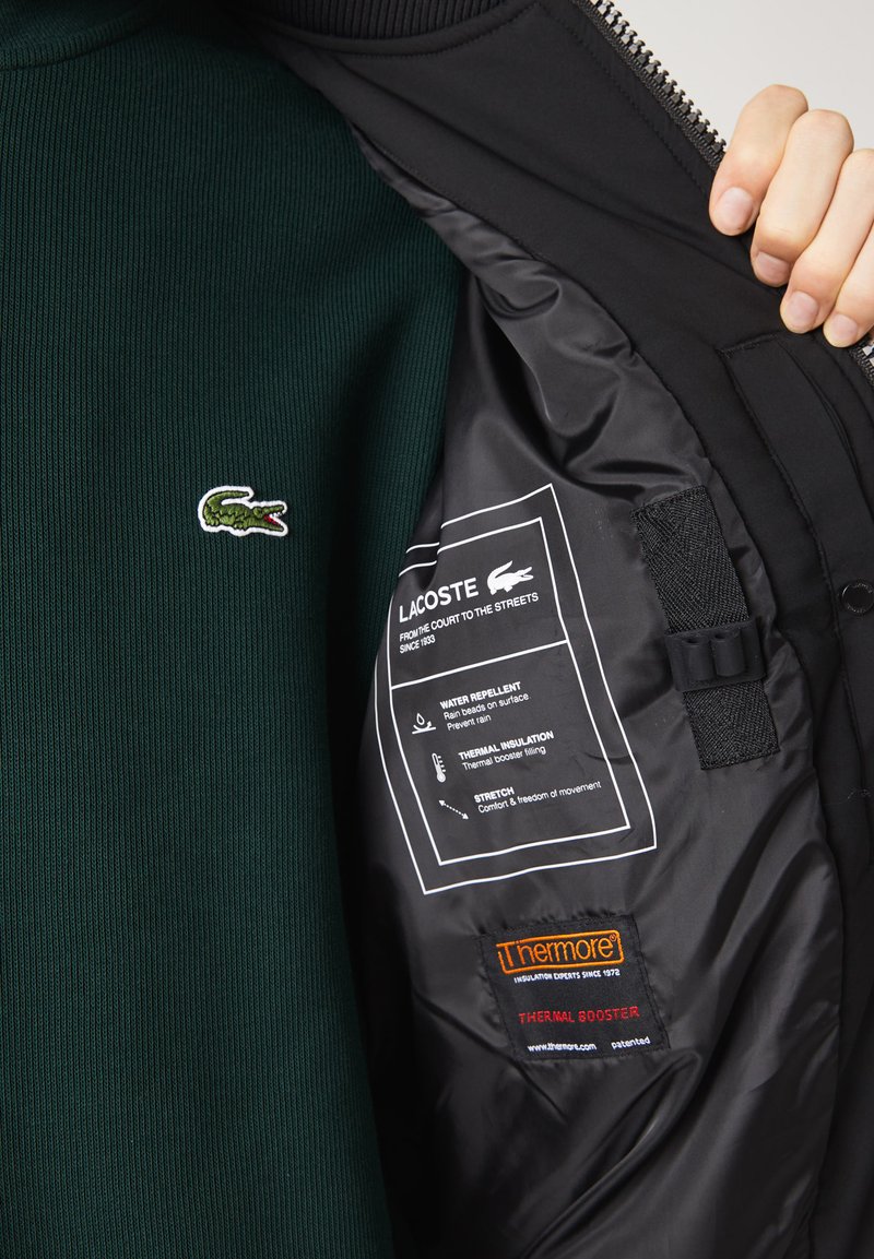 Pull en maille côtelée vert foncé avec un logo crocodile brodé blanc, à l'intérieur d'une veste noire montrant les étiquettes d'entretien et les détails d'isolation.