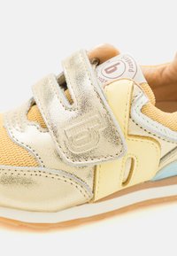 Sneakers dorés et jaunes avec une tige texturée, une sangle Velcro et une semelle rembourrée. Comprend un détail de marque sur la sangle et sur le côté.