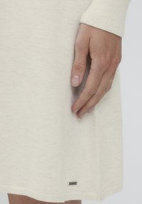 Rochie tricotată de culoare crem, cu manșete rigate, având un mic etichete. Textură moale, cu o formă în A și mici flecuri subtile pe întreaga țesătură.