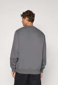Sudadera gris con cuello redondo, mangas largas y puños acanalados. Incluye un sutil ribete blanco a lo largo de los hombros. Textura de tela suave.