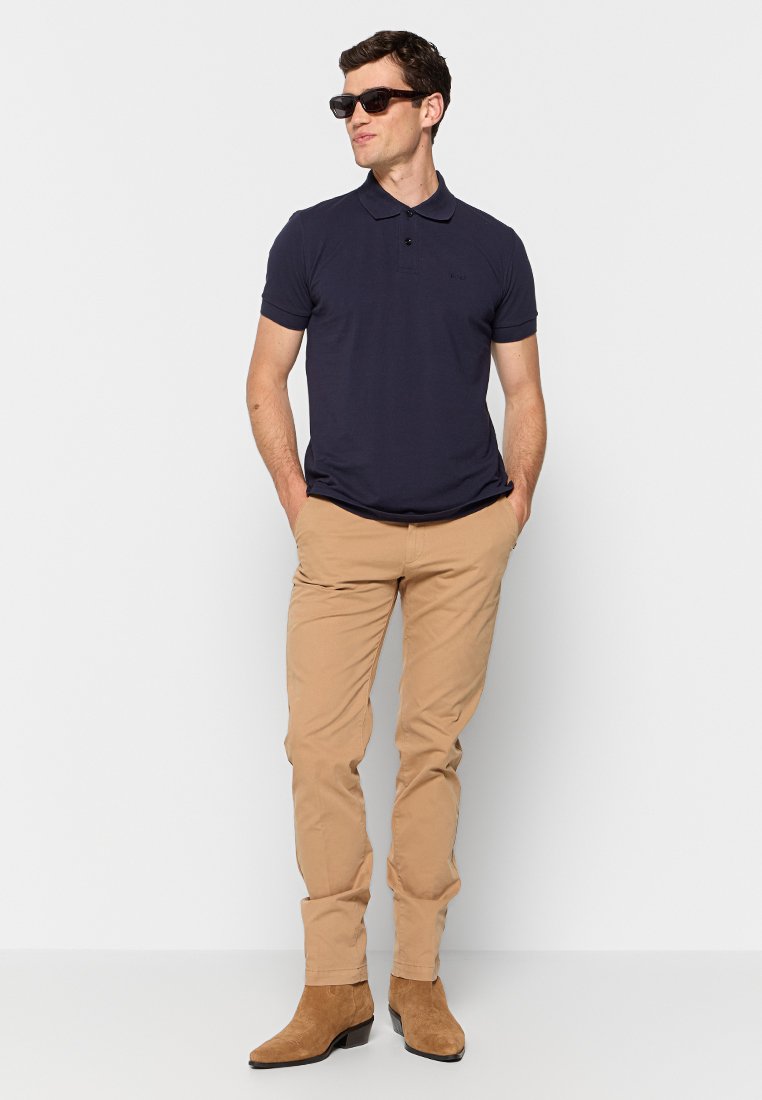 Polo shirt blu navy a maniche corte, pantaloni beige slim-fit e stivaletti alla caviglia in suede marrone. Il modello indossa occhiali da sole scuri e si trova in piedi con le mani in tasca.