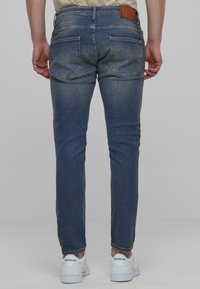 Tumed sinised denim teksad kitsase lõikega, taga taskutega ja pruunist nahast lipsuga vööl. Viimistletud kerge fade efektiga.