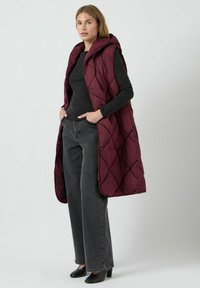 Mörkröd quiltad väst med huva, två knappar, bärs över en svart långärmad topp och grå vida byxor, tillsammans med svarta klackskor.