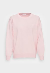 Różowy sweter z okrągłym dekoltem, długimi rękawami, ściągaczami przy rękawach i u dołu, z małym logo Lacoste na lewej piersi. Miękki materiał dzianinowy.