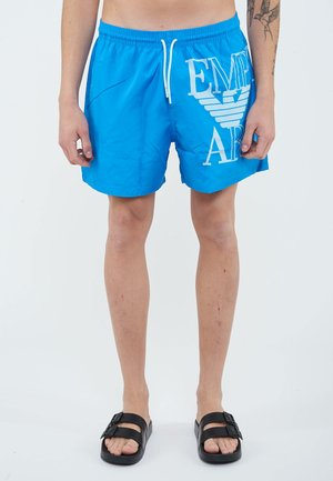 Uomo con pantaloncini da bagno blu brillante con logo bianco, sandali neri a ciabatta e un tatuaggio sul braccio sinistro, in piedi davanti a uno sfondo bianco.