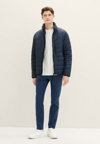 Marinblå puffjacka med ståkrage, dragkedjefickor och vadderad design, tillsammans med en ljusgrå långärmad tröja och marinblå byxor. Vita sneakers.