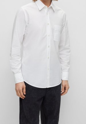Chemise blanche à manches longues, boutonnée, avec un col pointu et une poche poitrine gauche, fabriquée en tissu lisse et ayant un ourlet arrondi.