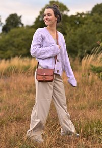 Cardigan en tricot cablé mauve clair avec de gros boutons, associé à un pantalon large beige et un sac à bandoulière en cuir marron. Fond vert.