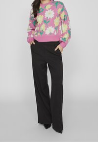 Person in schwarzen weiten Hosen und einem rosa Pullover mit großen gelben und weißen Blumenmustern, die Hände in den Taschen, vor einem grauen Hintergrund stehend.