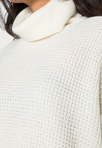 Suéter blanco de punto con un patrón texturizado y cuello tipo cowl. La tela parece suave y gruesa, adecuada para un uso casual en climas fríos.