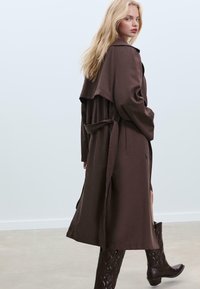 Bruine trenchcoat met een riem, brede revers en een vloeiende silhouet. Heeft een gestructureerde stof en is gecombineerd met donkere, gegraveerde cowboylaarzen.