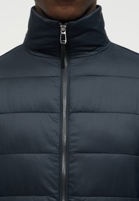 Κοντινό πλάνο ενός ατόμου που φοράει μπλε σκούρο puffer μπουφάν με ψηλό γιακά και ασημί φερμουάρ στο μπροστινό μέρος, κλειστό μέχρι το λαιμό.