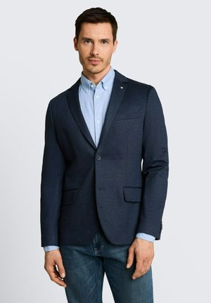 Homme portant un blazer bleu marine foncé sur une chemise bleu clair boutonnée et un jean bleu, debout devant un fond clair uni.