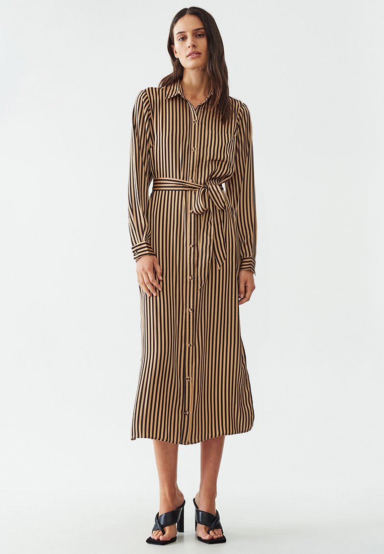 Hnědo-beige pruhované šaty s knoflíkovým zapínáním. Mají dlouhé rukávy, uvázaný pás a sahají do poloviny lýtek. Stylizované s podpatkovými sandály.