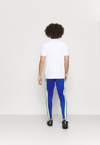 Person med lockigt hår som bär en vit kortärmad skjorta, blå och neongula träningsbyxor samt svarta sneakers, står vänd mot en plain vägg.