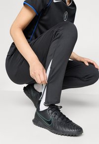 Sorte treningsbukser med blå detaljer, med glidelås på siden. Kombinert med sorte joggesko med grønne detaljer og hvite sokker.