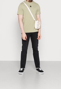 Olivgrön t-shirt med kort ärm och svart logotyp, svarta jeans, vita sneakers med svarta accenter, och en liten beige axelväska.