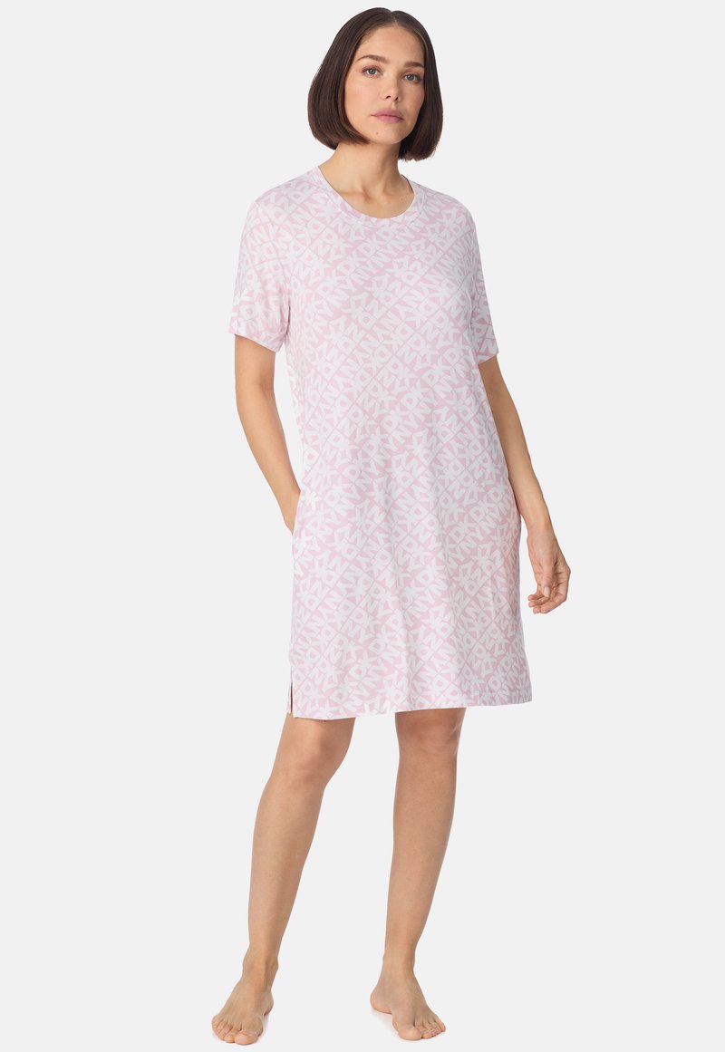 Femme debout, pieds nus, portant une robe rose clair mi-longue à manches courtes avec des motifs abstraits blancs, mains partiellement dans les poches.