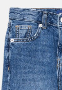 Nära bild av blå denimjeans som visar framfickan med nitar, bältes�öglan och kontrastsöm på ljus bakgrund.