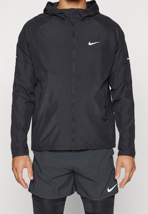 Mann trägt eine schwarze Nike-Kapuzenjacke mit Reißverschluss, schwarze Shorts und schwarze Leggings, steht vor einem einfarbigen Hintergrund.