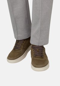 Baskets marron à lacets portées avec un pantalon gris clair, montrant les jambes inférieures et les pieds sur un fond uni.