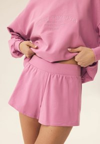 Jasnoróżowy sweter i dopasowane szorty, z prążkowanym paskiem w talii, miękkiego materiału oraz wytłoczonym tekstem na górze.