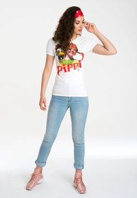 White cotton t-shirt with a colorful "Pippi" graphic print. Model wearing light blue denim jeans and pink sneakers, posed standing. Red bandana accessory.