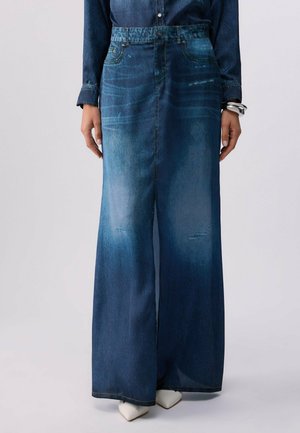 Mujer con pantalones vaqueros de pierna ancha y talle alto con lavado desgastado, combinados con una camisa vaquera y zapatos blancos de punta afilada.