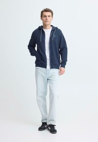 Sweat à capuche zippé bleu marine avec cordons contrastants, associé à un jean délavé clair et des baskets noires. Le sweat à capuche présente un petit logo sur la poitrine.