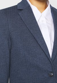 Blazer bleu marine à motif en chevrons, avec un col cranté, fermeture à un bouton et une chemise blanche à col en dessous.