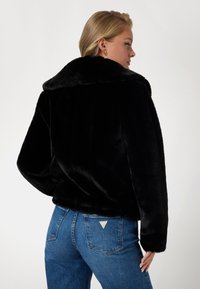Veste en fausse fourrure noire avec un large col, coupe courte et manches longues, associée à un jean bleu avec un patch triangulaire sur la poche arrière.