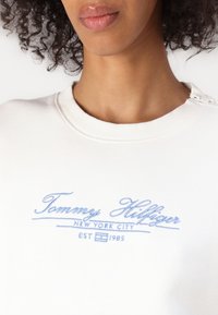 Hanorac alb cu guler rotund, cu tiv zimțat, logo-ul brodat "Tommy Hilfiger" în bleu deschis și detalii cu nasturi pe umăr.