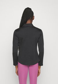 Zwarte langärmige blouse met een kraag en een gebogen zoom, gemaakt van een soepele stof. Gied gedragen over roze leggings, wat een slanke silhouet laat zien.