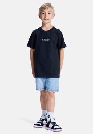 T-shirt en coton noir avec le logo "DC Shoes", associé à un short en jean bleu clair, des chaussettes blanches et des baskets noires avec des accents rouges.