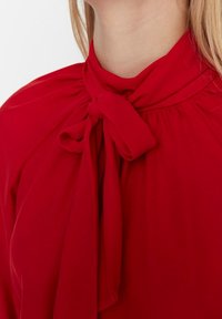 Red blouse with a tie at the neck, featuring a soft, smooth fabric, gathered detailing, and a relaxed fit around the shoulders.