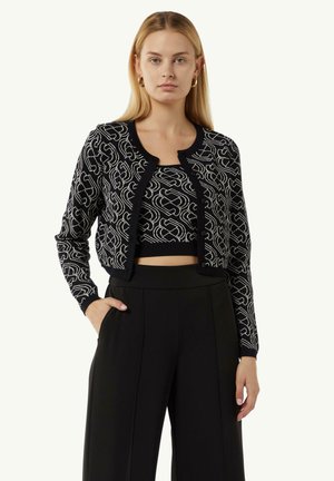 Schwarze, verkürzte Strickjacke mit weißem abstraktem Muster, Rundhalsausschnitt und Knopfverschluss. Getragen über einem passenden schwarzen Oberteil und taillierten schwarzen Hosen.