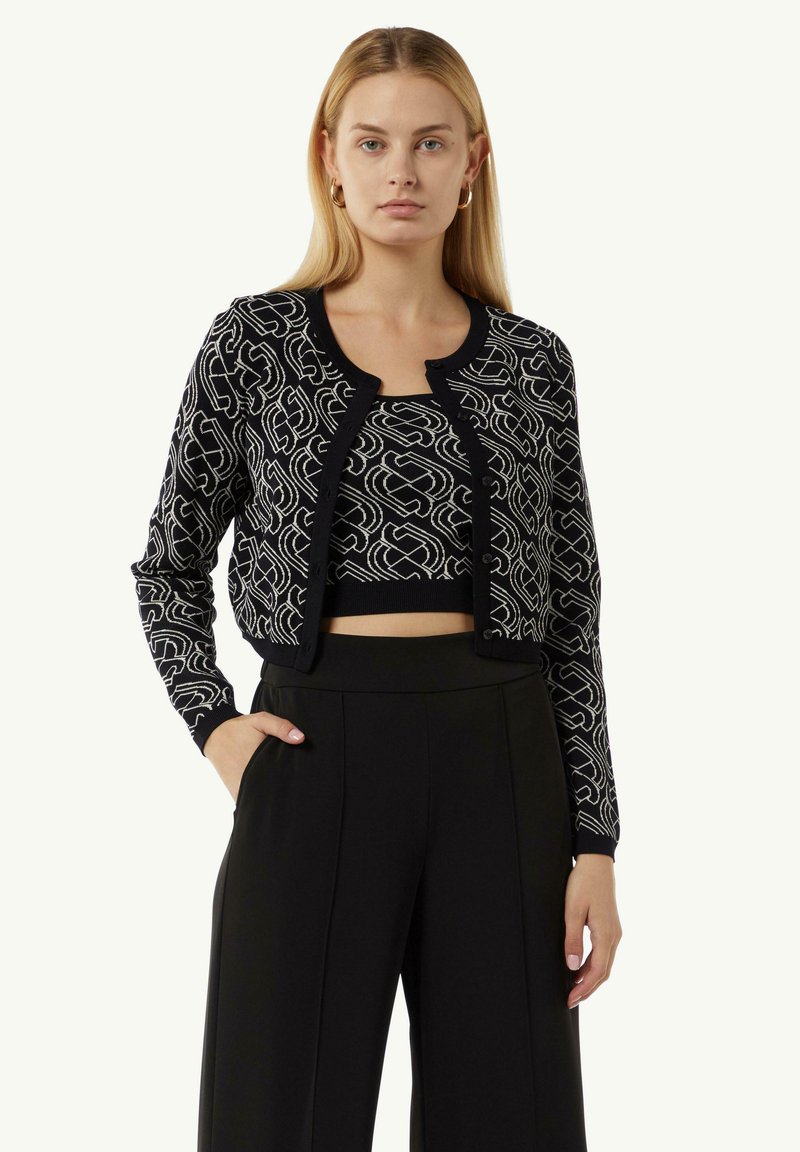 Schwarze, kurze Strickjacke mit weißem abstraktem Muster, strukturierter Strickstoff, Knopfverschluss und kontrastierendem Halsausschnitt. Getragen über einem schwarzen Oberteil und einer schwarzen Hose.