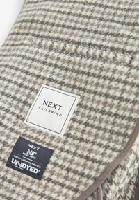 Uldblandingsstof i houndstooth-mønster med hvide og grå nuancer. Etiketterne inkluderer "NEXT Tailoring" og "UNDYED" med markante sorte accenter.