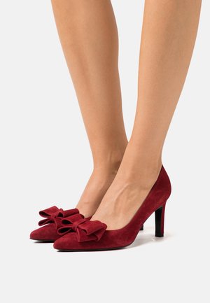 Tacchi alti in suede rosso con punta appuntita, caratterizzati da grandi fiocchi sul davanti e un sottile tacco nero. Texture liscia in tutto.