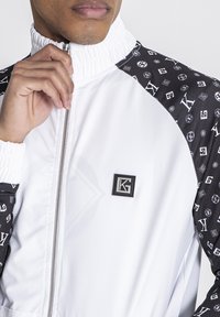 Chaqueta blanca con cremallera y cuello alto, con mangas de patrones negros. Parche con logo en el pecho. Tela suave, puños elásticos.