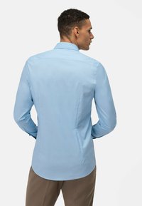 Chemise à manches longues bleu clair avec un col soigné, une texture lisse, des coutures discrètes et un ourlet incurvé à l'arrière.