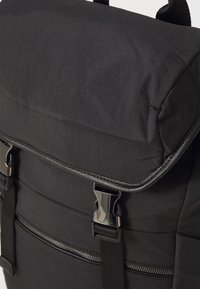 Zaino nero con una patta imbottita, dotato di una chiusura a fibbia lucida, tasca con zip e tessuto testurizzato per una maggiore durata.