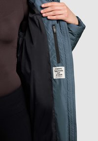 Die innere Auskleidung der blauen Steppjacke besteht aus schwarzem Stoff und verfügt über eine Reißverschlusstasche sowie ein Etikett mit der Aufschrift "Eine warme Umarmung aus Stoff."