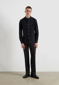 Schwarzes Hemd mit Knöpfen in Weiß und einem kleinen orangefarbenen Logo, kombiniert mit dunklen, maßgeschneiderten Hosen und schwarzen Schuhen. Glatte Stofftextur.