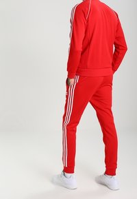 Röd träningsdräkt i glattyg, med vita ränder längs ärmarna och sidorna. Vita sportskor fulländar looken.