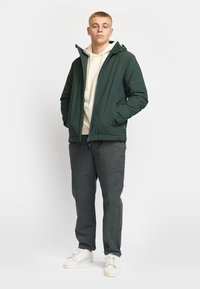 Dark green insulated jacket with a hood, paired with a light hoodie and dark gray pants. Model wears white sneakers.