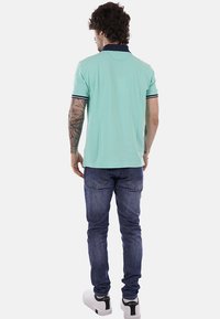 Homme tatoué portant un polo turquoise clair avec un col bleu marine, un jean bleu foncé et des baskets blanches, debout de dos sur un fond blanc.