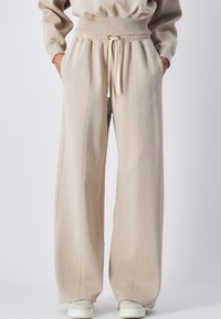 Pantaloni della tuta larghi beige realizzati in morbido misto cotone, con vita elastica e cordoncino, tasche laterali e dettagli cuciti.