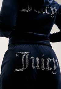 Navy žametni trenirni komplet z okrasnimi kamenčki in pisanim napisom "Juicy" na hrbtu, elastičnim pasom in oprijetim dizajnom.