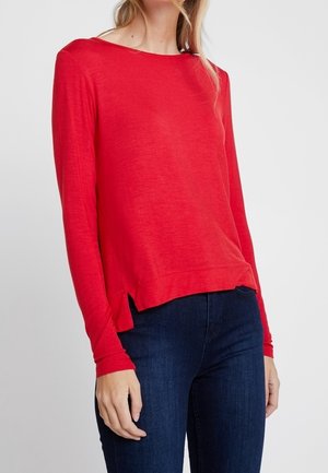 Femme vêtue d’un pull rouge à manches longues et d’un jean bleu foncé sur un fond blanc uni.